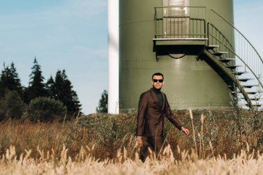 Yeşil golf tişörtlü bir iş adamı, yel değirmeninin yanında, arka planda ve mavi gökyüzünde duruyor. Yel değirmenlerinin yakınında bir iş adamı.