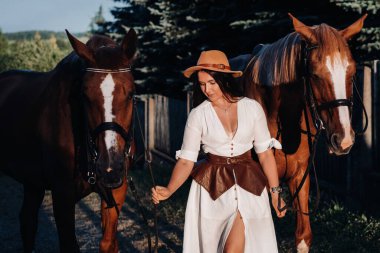 Beyaz elbiseli ve şapkalı bir kız kartal ahırında atların yanında duruyor.