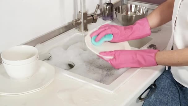 Μια γυναίκα με ροζ πλαστικά γάντια αφρού πλένει ένα πιάτο σε ένα μεγάλο νεροχύτη στο σπίτι — Αρχείο Βίντεο