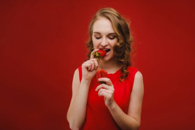 Kırmızı arka planda kırmızı elbiseli güzel bir kız elinde bir çilek tutuyor ve gülümsüyor..