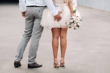 The groom puts his hand on the ass of his bride. The bride in a short dress back holding a bouquet in her hands.