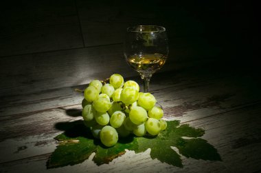 Sonbahar şarap yapma zamanıdır. Bir bardak şarap, bir demet beyaz üzüm ve ahşap zemin üzerinde üzüm yaprakları ile kompozisyon.