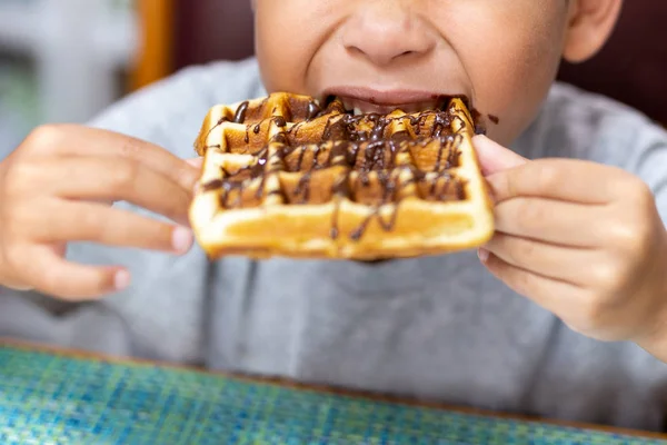 Dítě bitting oplatka s čokoládovou polevou u stolu se snídaní. — Stock fotografie