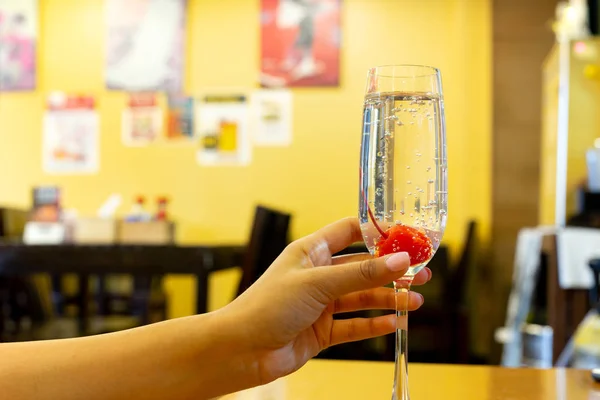 Mulher mão segurando copo de champanhe com cereja no restaurante com parede amarela . — Fotografia de Stock