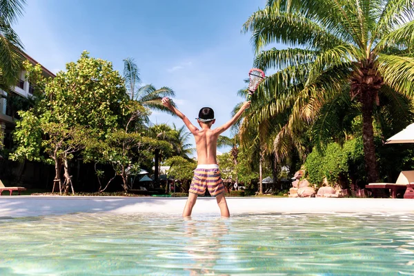 Mão menino feliz chegando e segurando bola e raquete de pé na piscina em férias . — Fotografia de Stock
