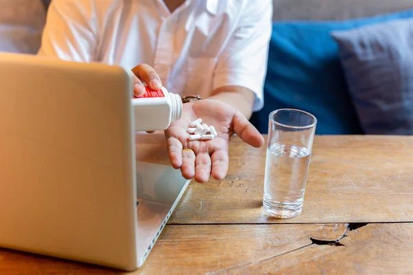 Man taking pill with glass of water and laptop on wooden table