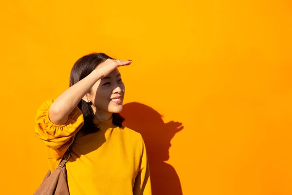 Asian woman  smiling in orange top with hand on forehead looking at camera in orange background