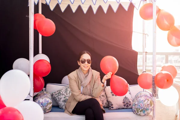 Woman with suit and scarf sitting on a couch hand holding red balloons