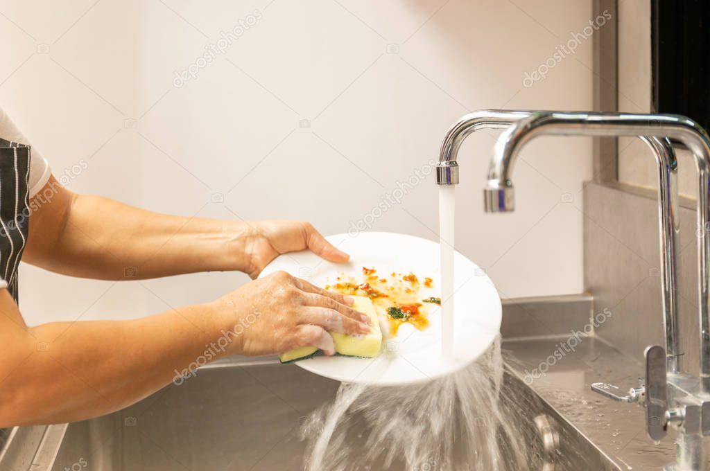 Hands washing dirty dishes with running water in kitchen sink