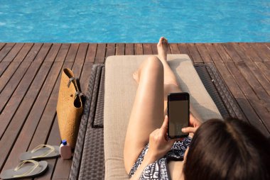 Güzel bayan plaj çantası ile sandalye yüzme havuzunun kenarında yatarken cep telefonu kullanarak.