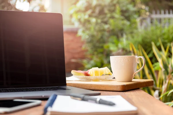 Kaffeebecher mit frischem Obst und Laptop auf Holztisch im Garten. — Stockfoto