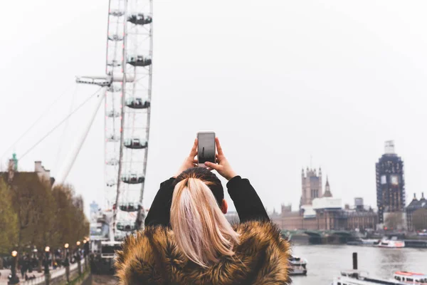 Tourist woman taking photo of London Eye with cell phone by thames river . — Fotografia de Stock