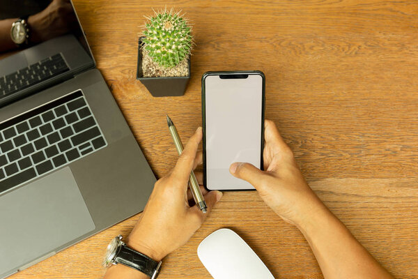 Businessman hand using cell phone and holding pen with laptop on wooden desk.