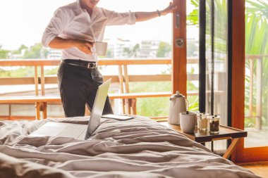 Otel odasında kahve tutan işadamı standı ile yatakta açık dizüstü bilgisayar.