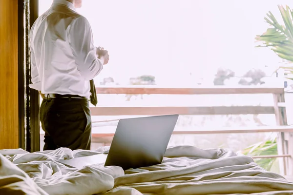 Laptop on the bed with businessman holding cup of coffee looking out of hotel room.