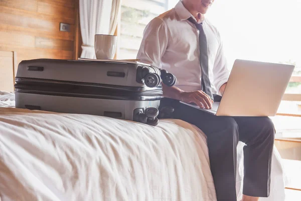 Επιχειρηματίας κάθεται στο κρεβάτι δουλεύοντας σε φορητό υπολογιστή με κούπα καφέ πάνω από τη βαλίτσα. — Φωτογραφία Αρχείου