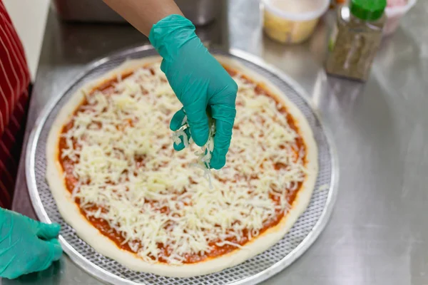 Main de cuisinier avec des gants verts mettant du fromage sur la pizza crue . — Photo