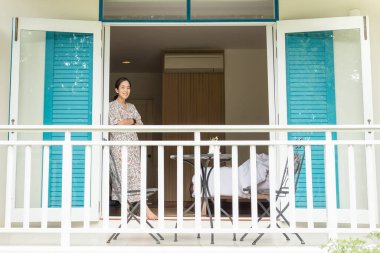 Açık pencerenin yanında duran turist kadın otel odasının güzel manzarasına bakıyor.