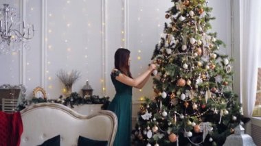 Koyu yeşil elbiseli oldukça uzun saçlı bir kadın lüks iç mekandaki Noel ağacını süslüyor.. 