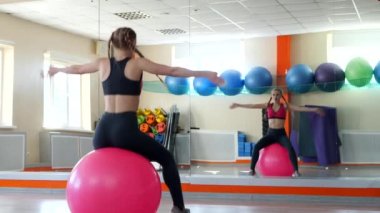 Pembe spor bluzlu, uzun saçlı ve siyah taytlı güzel bir kız pembe lastik topun üzerinde jimnastik yapıyor. Zıplayarak ve topun üstüne çömelerek eğleniyor..
