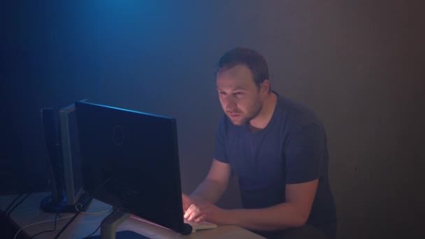 男性黑客正在认真地完成他在计算机上的工作 它会发出恶意蠕虫来窃取用户的个人资料 蓝色的霓虹灯照亮了一间黑暗的办公室 桌上有两个监视器 — 图库视频影像