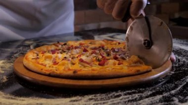 Şef özel bir bıçakla pizzayı karideslerle birkaç parçaya ayırıyor. Eldivenli ellerin yakın çekimi. Üzerinde un olan siyah bir masada pizza..