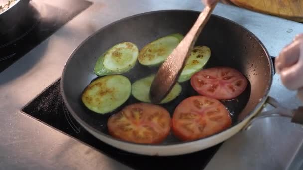 Chef is frying vegetables in a large frying pan. Eggplant, zucchini, grilled tomatoes are fried in oil. Cook turns the ingredients with a wooden spatula.