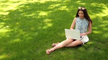 Çekici kadın dizüstü bilgisayarında yazıyor, internetten iletişim kuruyor, sohbet ediyor ve elini sallıyor. Güzel esmer kız parkta çimlerin üzerinde oturuyor. Yeşil arkaplan, kopyalama alanı.