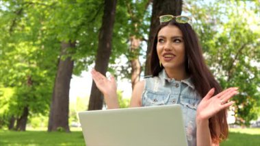 Uzun saçlı çekici kız parkta dizüstü bilgisayarda web kamerasıyla iletişim kuruyor. Konsept: Serbest çalışmak, ofis dışında çalışmak. Kadın kameranın önünde duygusal jestler yapıyor..