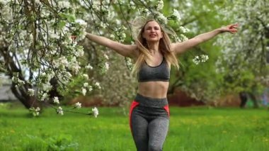 Sportif kadın baharda parkta kardiyo egzersizleri yapıyor. Kadın zıplıyor ve derin nefes alıyor. Parkta üç tane çiçek açmıştır. Kız üst ve tayt giyiyor..