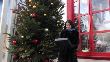 Kürk manto giymiş çekici bir kadın, üzerinde mutlu yıllar yazan bir tablet gösteriyor. Arka planda tatil vitrini ve Noel ağacı var. Aralık 'ta dışarıda..