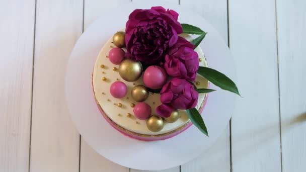 ペストリーボールと新鮮な花で飾られた誕生日ケーキは 木製の背景に白いスタンドに円で回転します 上からの眺め — ストック動画