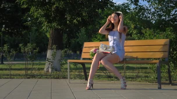 Μια όμορφη κοπέλα ακούει μουσική στα ακουστικά της ενώ κάθεται στο πάρκο. — Αρχείο Βίντεο