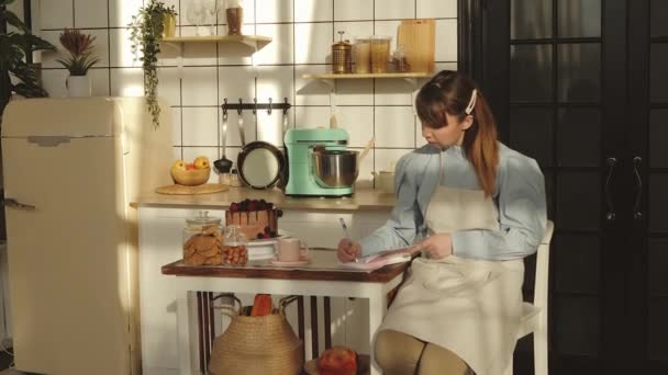 Beautiful woman is making notes while cooking in the kitchen. — Stock Video