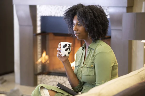 暖炉のソファでくつろぎながら 自宅でコーヒーやCbdティーを飲みながら休んでいる黒人アフリカ系アメリカ人の女性 彼女はリビングルームで暖かいカップを持っている — ストック写真