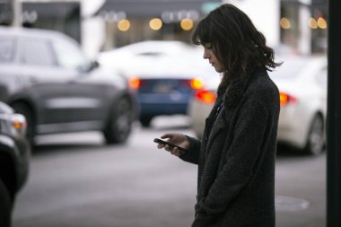Female pedestrian waiting on a sidewalk for a rideshare.  She is sharing her gps location via cellphone app so the driver can pick her up in the city.  Cars are blurred to obscure make model and license plates.  clipart