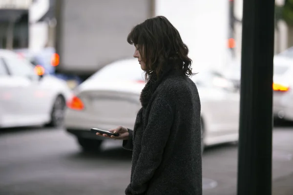 Female pedestrian waiting on a sidewalk for a rideshare.  She is sharing her gps location via cellphone app so the driver can pick her up in the city.  Cars are blurred to obscure make model and license plates.