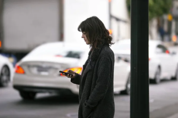 Female pedestrian waiting on a sidewalk for a rideshare.  She is sharing her gps location via cellphone app so the driver can pick her up in the city.  Cars are blurred to obscure make model and license plates.