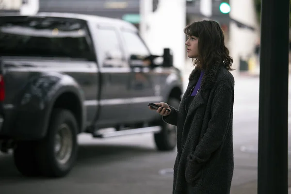 Female pedestrian waiting on a sidewalk for a rideshare.  She is sharing her gps location via cellphone app so the driver can pick her up in the city.  Cars are blurred to obscure make model and license plates.