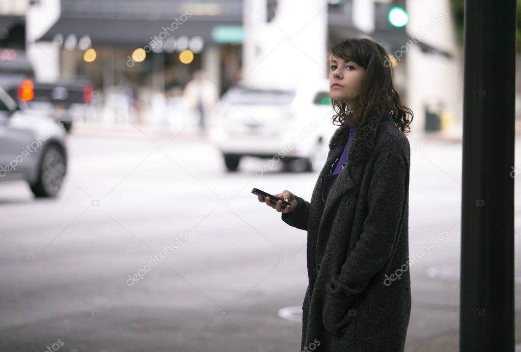 Female pedestrian waiting on a sidewalk for a rideshare.  She is sharing her gps location via cellphone app so the driver can pick her up in the city.  Cars are blurred to obscure make model and license plates. 