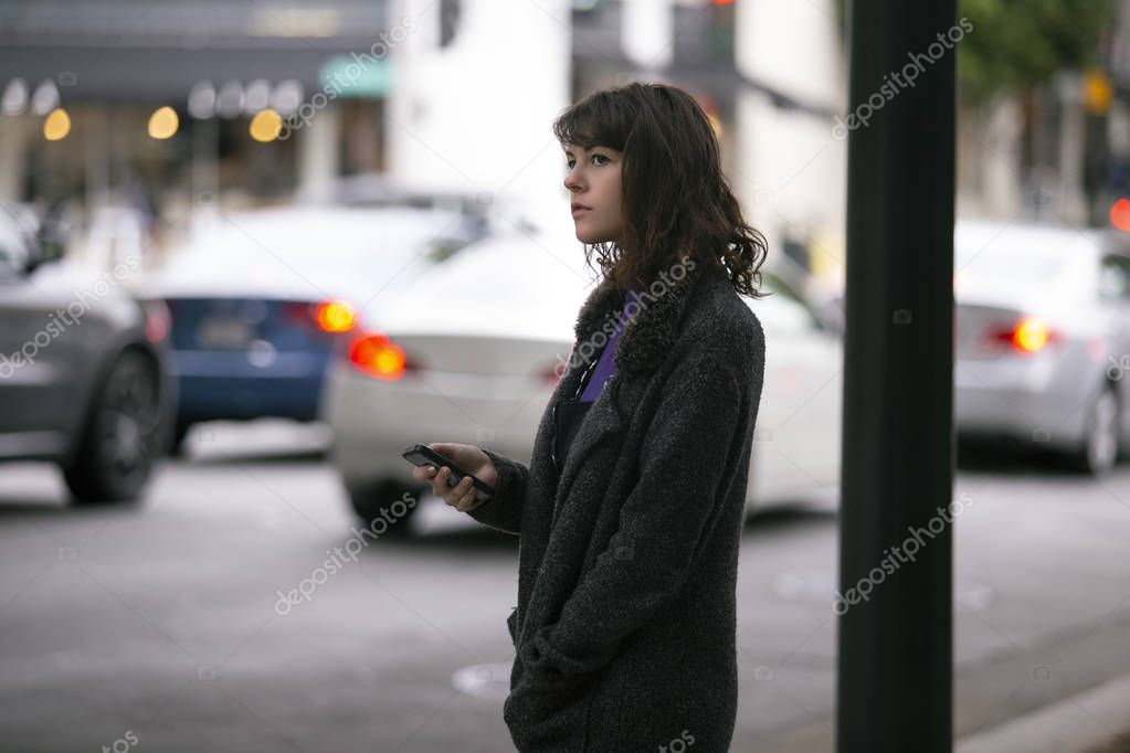 Female pedestrian waiting on a sidewalk for a rideshare.  She is sharing her gps location via cellphone app so the driver can pick her up in the city.  Cars are blurred to obscure make model and license plates. 