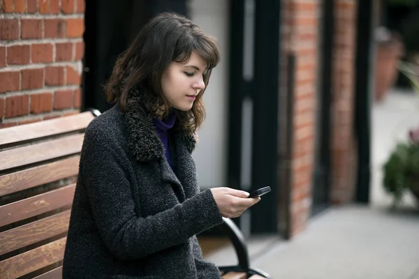 Young Caucasian female commuter at a bus stop or train station or a rideshare.  She is sitting on a bench and waiting patiently.  She is browsing on her cell phone.