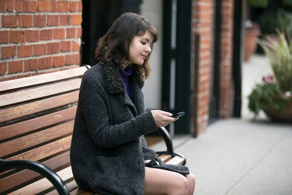 Young Caucasian female commuter at a bus stop or train station or a rideshare.  She is sitting on a bench and waiting patiently.  She is browsing on her cell phone.