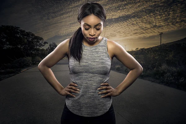 Athletic black African American female runner or jogger resting to pose with a motivated facial expression with a road during sunset in the background.  Depicts endurance and determination during marathon sports.