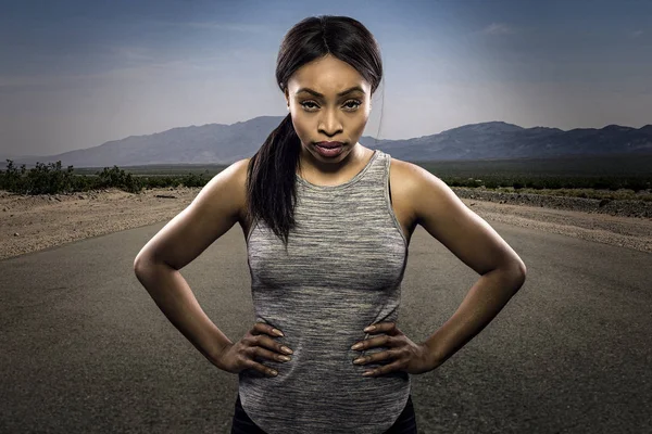 Athletic black African American female runner or jogger resting to pose with a motivated facial expression with a road during sunset in the background.  Depicts endurance and determination during marathon sports.