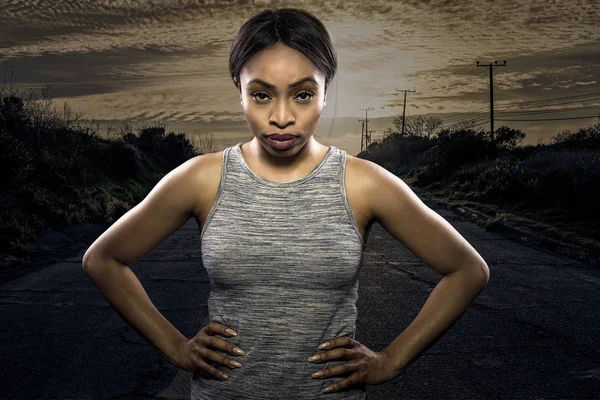 Athletic black African American female runner or jogger resting to pose with a motivated facial expression with a road during sunset in the background.  Depicts endurance and determination during marathon sports.