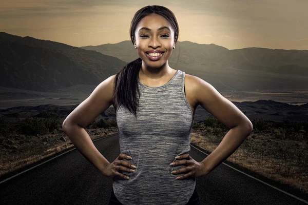Athletic black African American female runner or jogger resting to pose with a motivated facial expression with a road during sunset in the background.  Depicts endurance and determination during marathon sports.