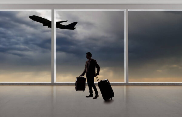 Silhouette of a traveling businessman with luggage in front of an airport window with a view of the sky and an airplane.  The panorama has copy space for text.  Depicts business travel.  