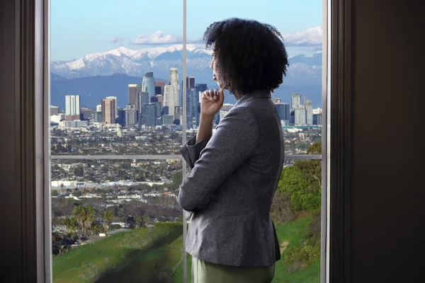 ロサンゼルスのオフィスの窓から外を見る黒人女性ビジネスウーマン 彼女は都市開発を考えている女性建築家のように見えるか 市長や知事がゾーニング法を計画しているように見えます — ストック写真