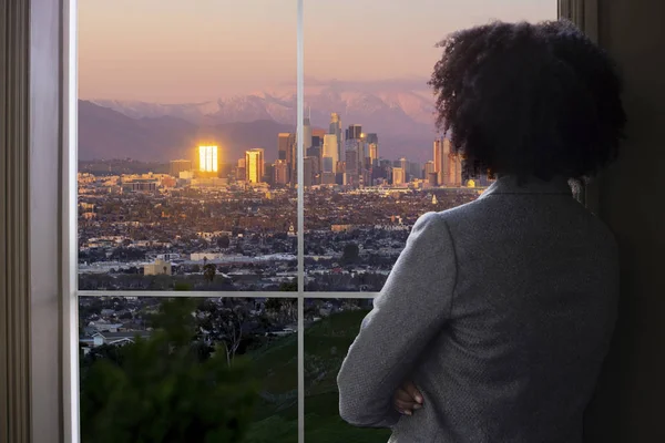 Black female business woman looking out the window of an office in Los Angeles.  She looks like a female architect thinking of urban development or a city mayor or governor planning zoning laws.